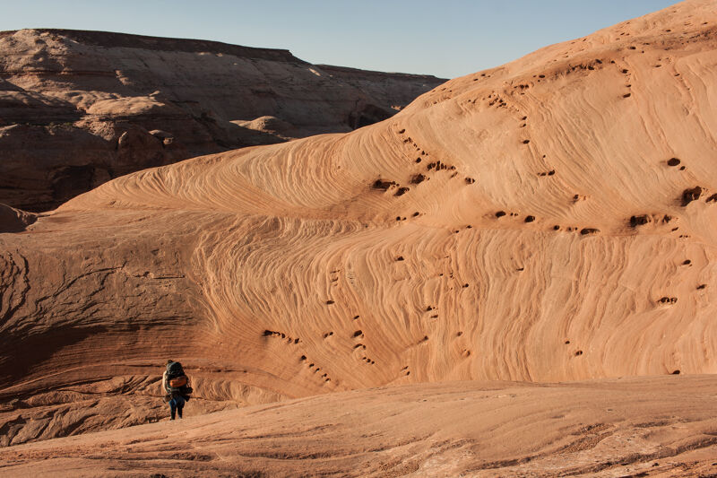 The image shows a desert landscape with a person hiking. The terrain is characterized by undulating sandstone formations, displaying layered patterns and textures. The color palette is dominated by warm earth tones, primarily shades of orange and brown. The sky is clear and light blue. The hiker is small in comparison to the vastness of the landscape, emphasizing the scale and grandeur of the natural environment.